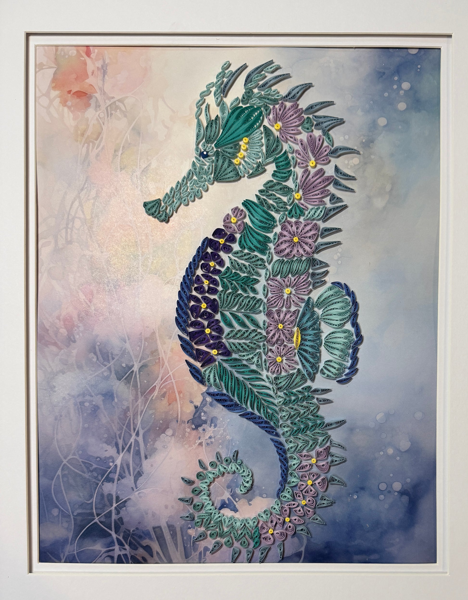 A framed art print featuring a stylized seahorse composed of layered green leaves and purple flowers, set against a soft blue and white watercolor background.